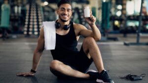 Young Muscular Arab Man Holding Container with Supplement Pills, Posing at Gym