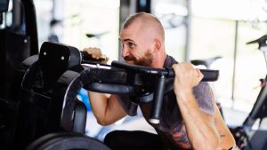 Free photo fit tattooed bearded man doing squats in a training machine