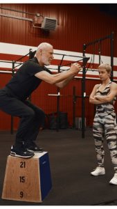 vertical-shot-beautiful-confident-woman-fitness-instructor-crossing-arms-her-chest-watching-her-elderly-male-doing-squats-building-up-muscles-during-personal-workout-modern-gym.